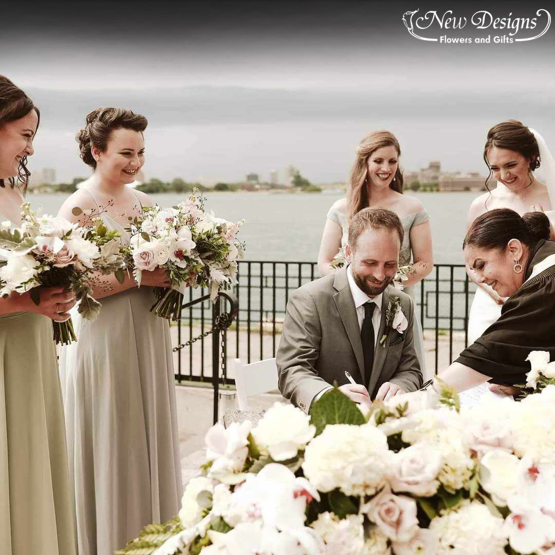 a bride and groom sign their registry on a table overflowing with roses and orchids as the bridesmaids stand to the side holding bouquets of lush roses and Phalaenopsis orchids.
